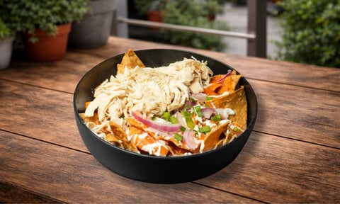 Black bowl of chilaquiles topped with creamy sauce, tortilla chips, red onion, and cheese on wooden table with garden backdrop