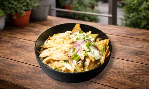 Bowl negro de papas fritas con queso, bacon y cebollín sobre mesa de madera con plantas en maceta