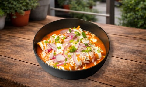 A bowl of vibrant orange curry soup topped with fresh herbs, pink onions, and white garnish on a wooden outdoor table