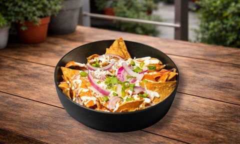 Nacho nachos topped with cheese, sour cream, jalapeños, and green onions served in a black bowl on an outdoor wooden table