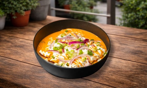 Black bowl of creamy orange curry soup with vegetables and herbs on a wooden outdoor table