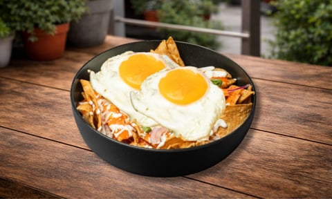 Bowl negro con chilaquiles y dos huevos fritos sobre mesa de madera en patio exterior