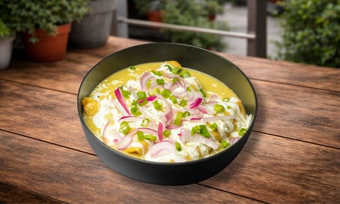 Bowl of creamy soup with corn, pink onions, and green herbs on a wooden table with potted plants in background